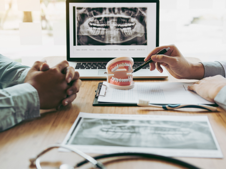 A dental consultation setting with a dentist and a patient discussing a treatment plan. The dentist is holding a pen and pointing at a document, while the patient has their hands clasped together. On the table, there is a clipboard with a dental X-ray image, a laptop displaying the same X-ray, a dental model, and a toothbrush.
