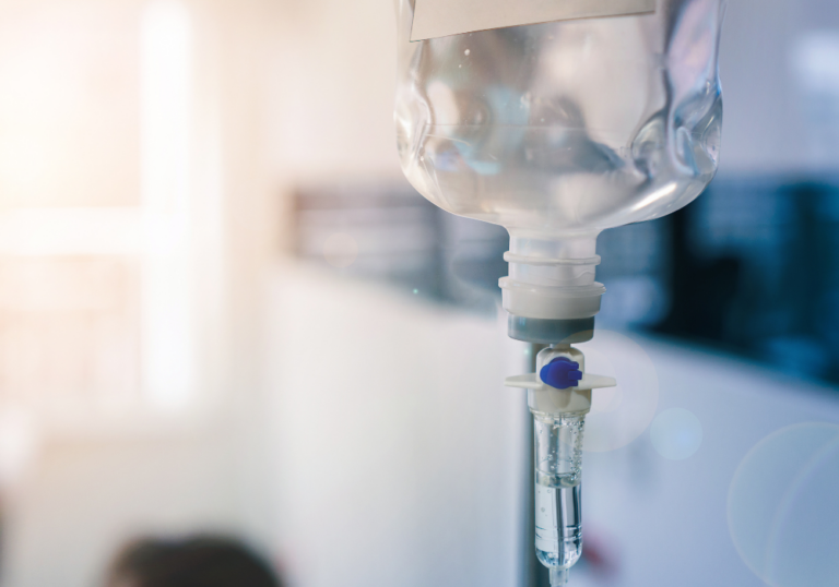 Intravenous (IV) drip bag partially filled with clear liquid, connected to a drip chamber and tubing, in a clinical environment.